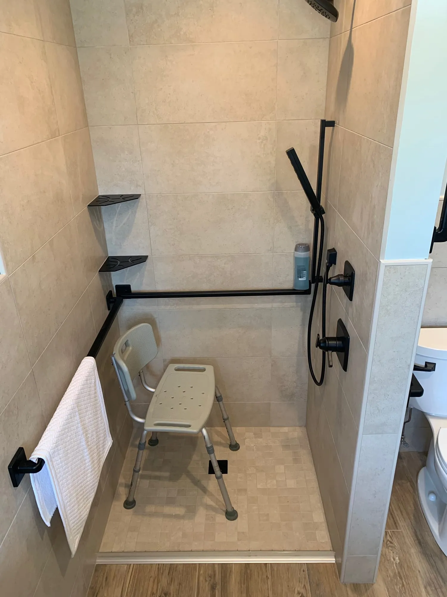 Accessible shower viewed from the bathroom, with grab bars, corner shelves and a shower chair