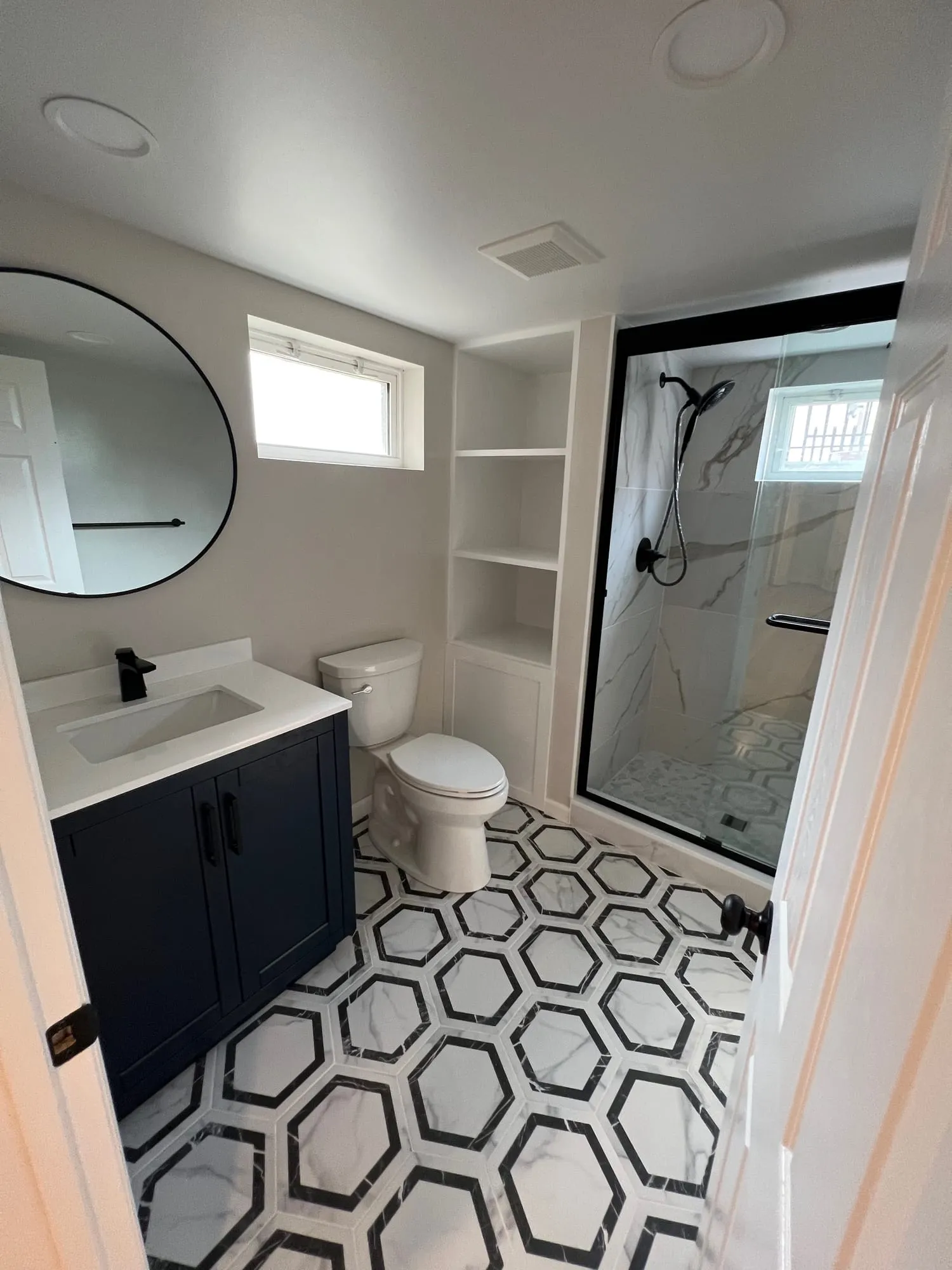 Basement bathroom with black-and-white hexagon floor tile, a navy vanity, round mirror and glass shower