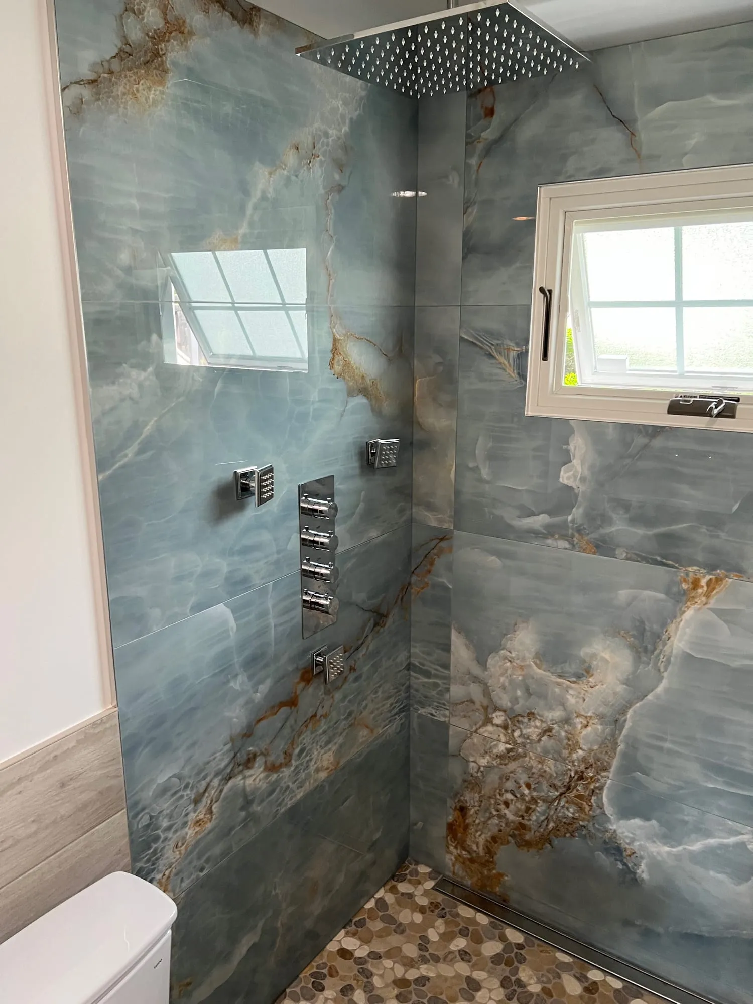 Nesconset shower with blue onyx-look porcelain walls, rain head, body sprays and a pebble floor
