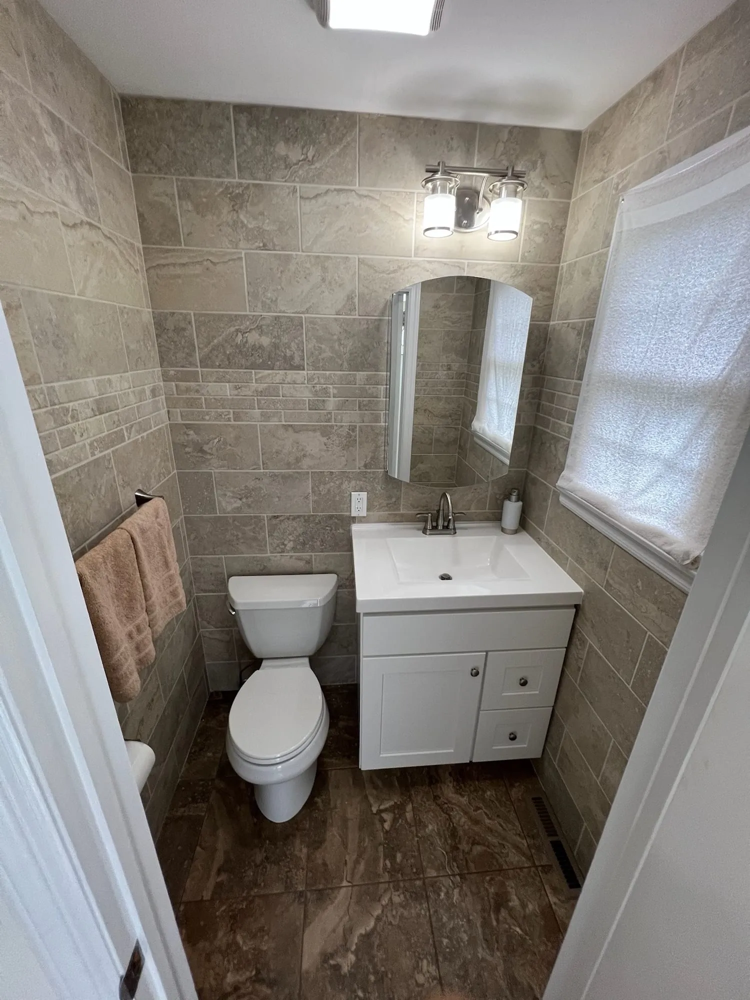 Half bath with stone-look wall tile, a white vanity, wall sconce and dark marble-look floor tile