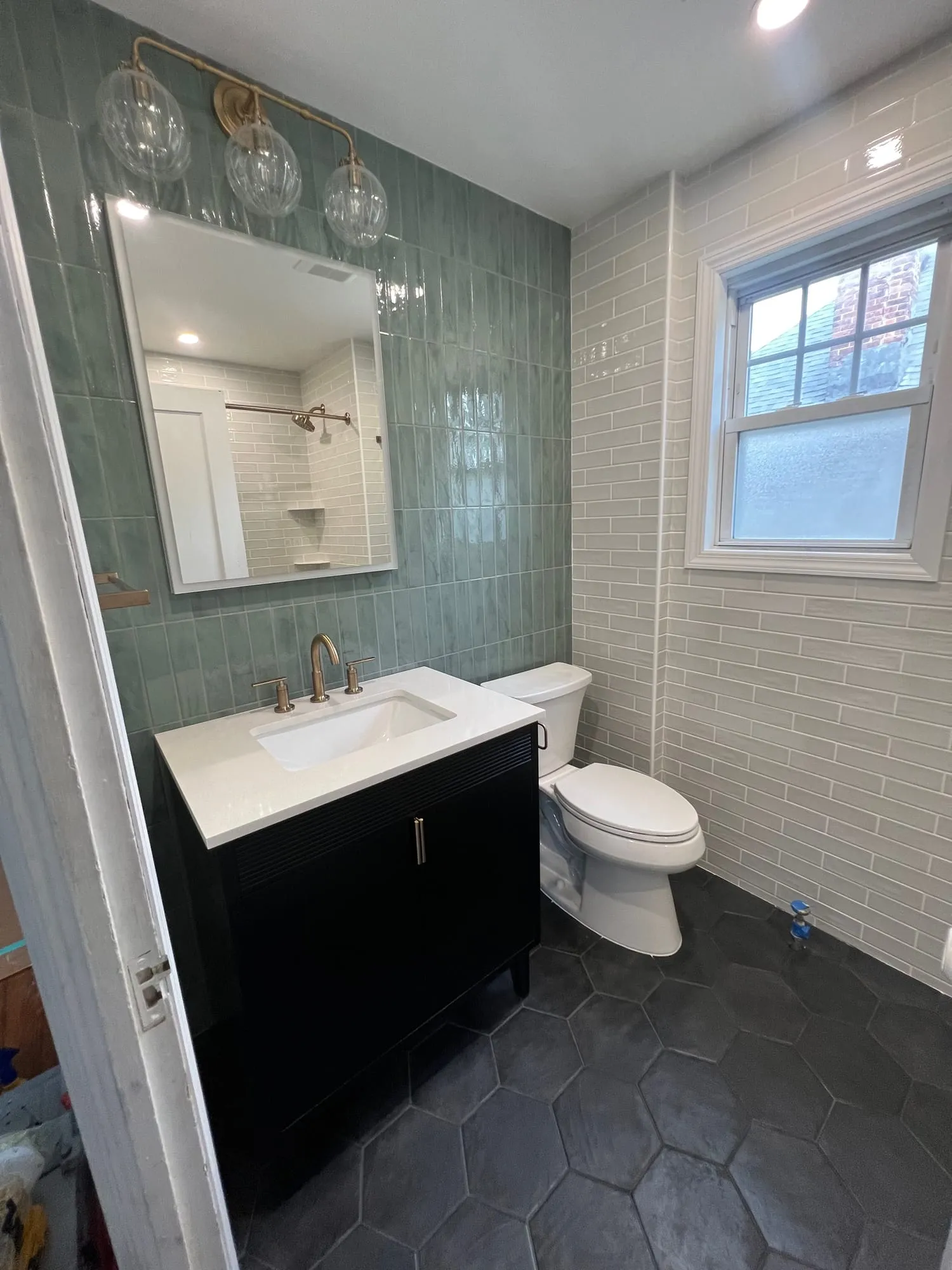 Bathroom with a sage glazed tile accent wall, brass fixtures, black vanity and black hexagon floor tile