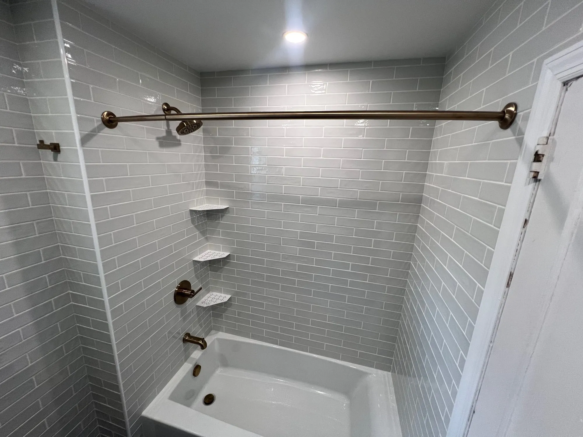 Tub surround in light gray subway tile with brass shower fixtures and corner shelves